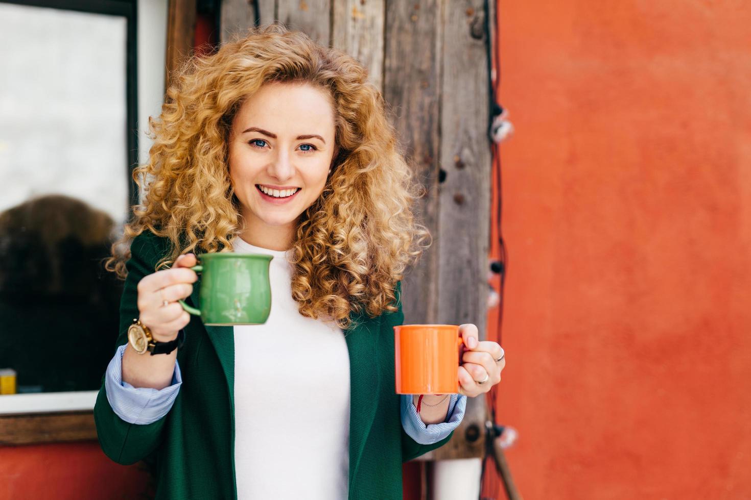 femme heureuse aux cheveux blonds et bouclés charmants yeux bleus tenant deux tasses de café va traiter ses amis avec du café ayant une expression heureuse et un sourire sur son visage. notion de langage corporel photo