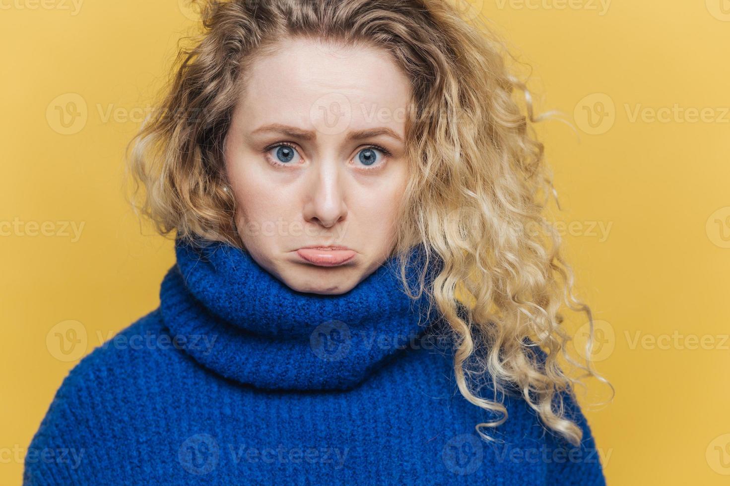 séduisante jeune femme offensée a les cheveux bouclés et touffus, courbe la lèvre inférieure en signe d'insatisfaction, se sent bouleversée après une querelle avec son mari, porte un pull bleu, isolée sur fond de studio jaune. photo