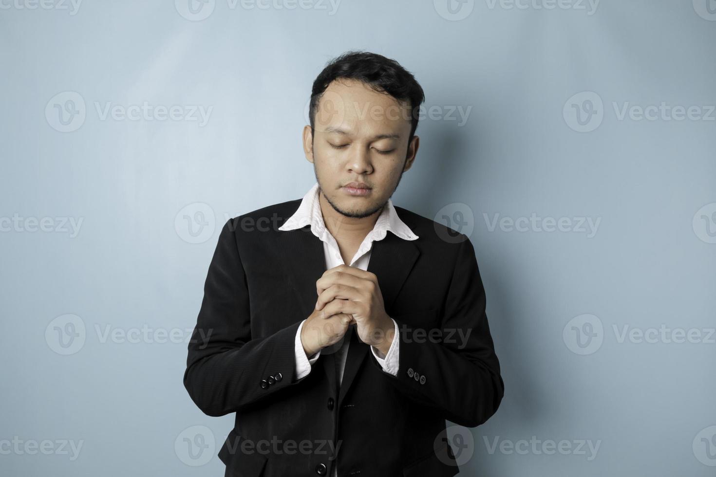 un portrait d'un homme d'affaires asiatique portant un costume noir prie pour le succès isolé par un arrière-plan bleu photo