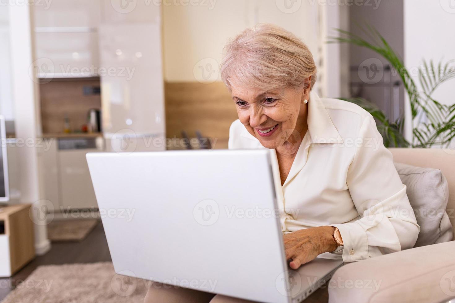 portrait d'une femme âgée assise à table à la maison et travaillant sur son ordinateur portable. dame âgée surfant sur le net depuis la maison assise sur son canapé et utilisant un ordinateur portable photo