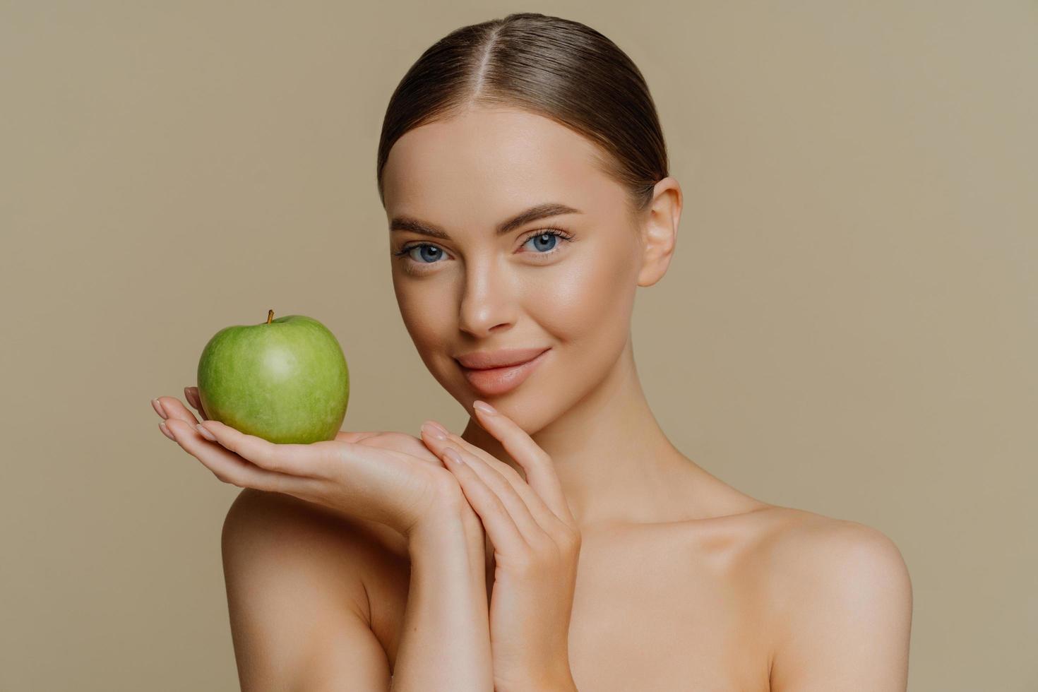 photo d'une jeune femme brune torse nu aux cheveux peignés tenant une pomme verte fraîche utilise des cosmétiques naturels a des épaules nues isolées sur fond marron. concept de soins du corps cosmétologie personnes