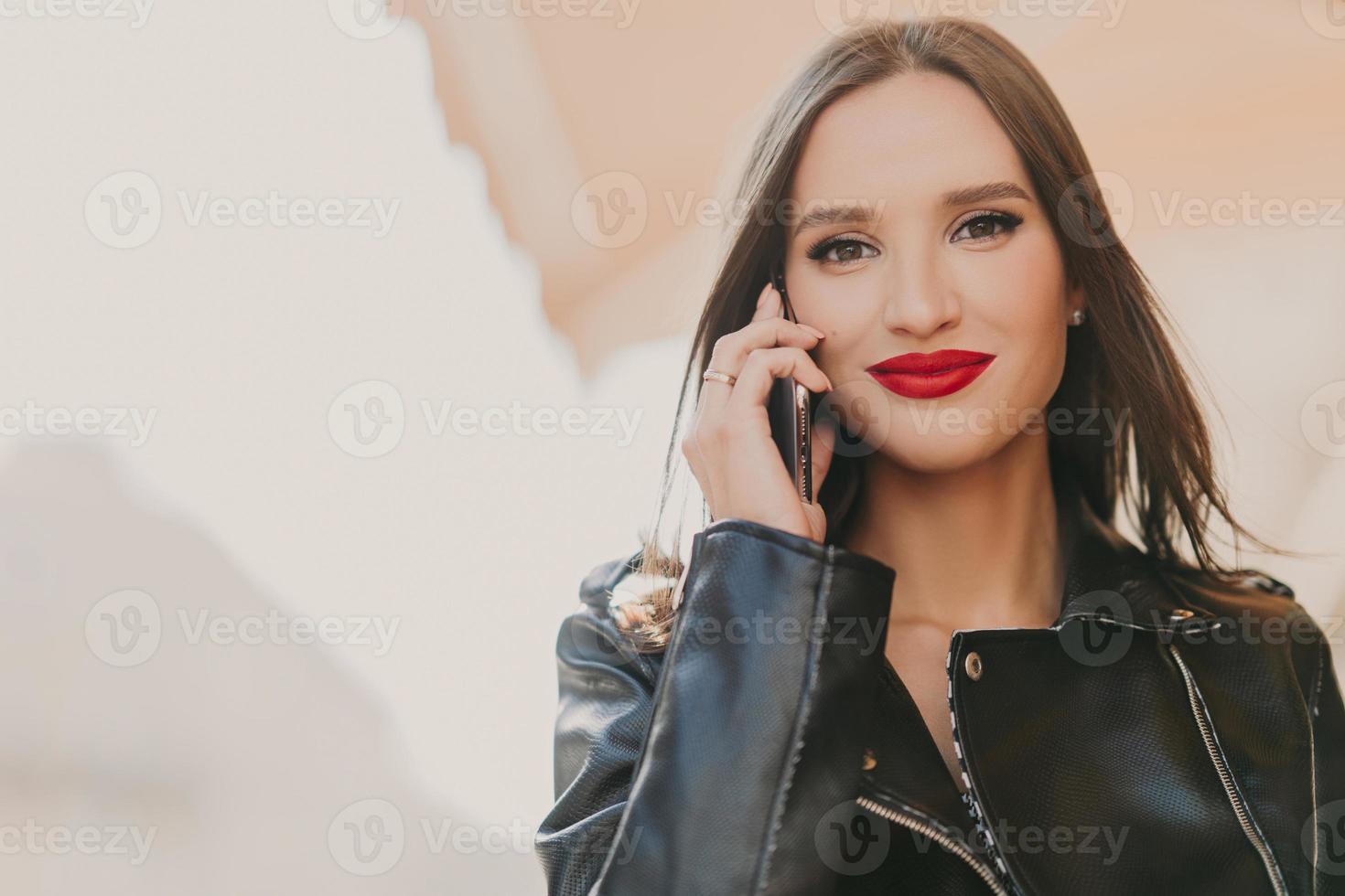 une jolie femme séduisante bénéficie de tarifs en itinérance, parle au téléphone portable avec son meilleur ami, vêtue de vêtements à la mode, porte du rouge à lèvres, se maquille, se tient sur l'arrière-plan flou d'un bâtiment photo