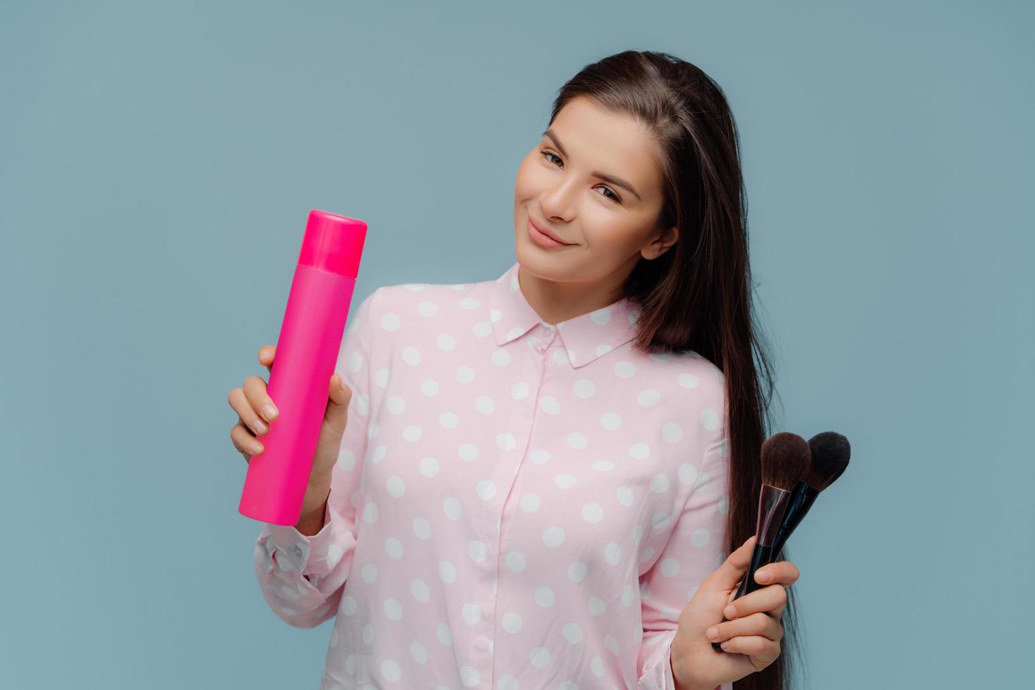 une femme brune aux cheveux longs satisfaite utilise de la laque pour faire une coiffure élégante, des pinceaux cosmétiques pour appliquer de la poudre sur le visage, se prépare pour une occasion spéciale, porte une chemise élégante, isolée sur un mur bleu photo