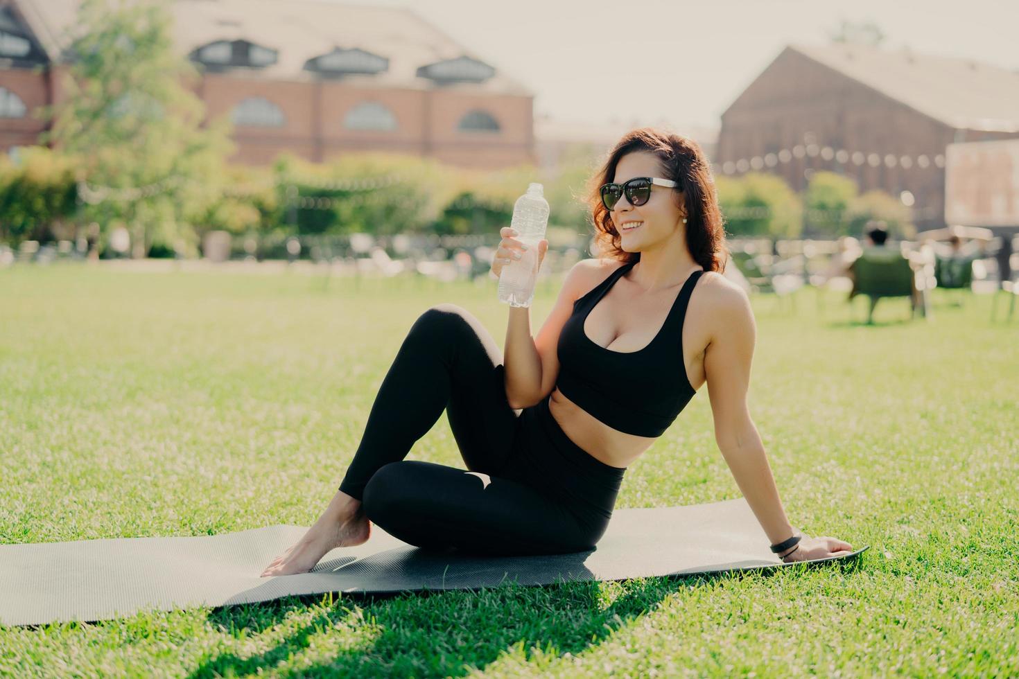 heureuse jeune femme européenne sportive se repose après l'entraînement pose sur un tapis de fitness avec les pieds nus boit de l'eau vêtue de lunettes de soleil actives mène un mode de vie sain. concept d'hydratation d'entraînement des personnes photo