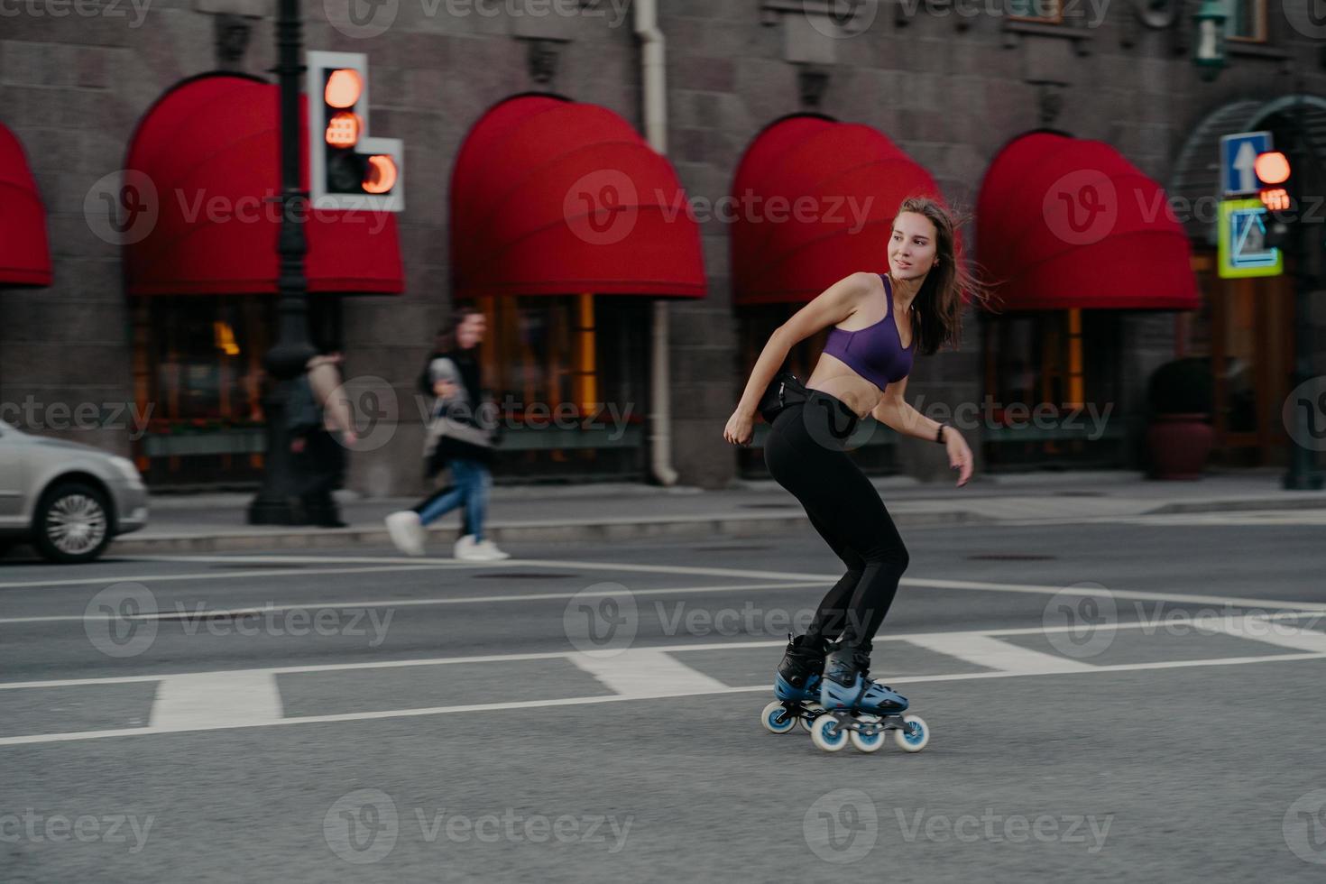 une jeune femme sportive et mince utilise des patins à roulettes comme mode de transport en ville aime son passe-temps préféré et améliore son humeur vêtue de vêtements de sport pose sur la route s'exerce régulièrement garde la forme perd du poids photo