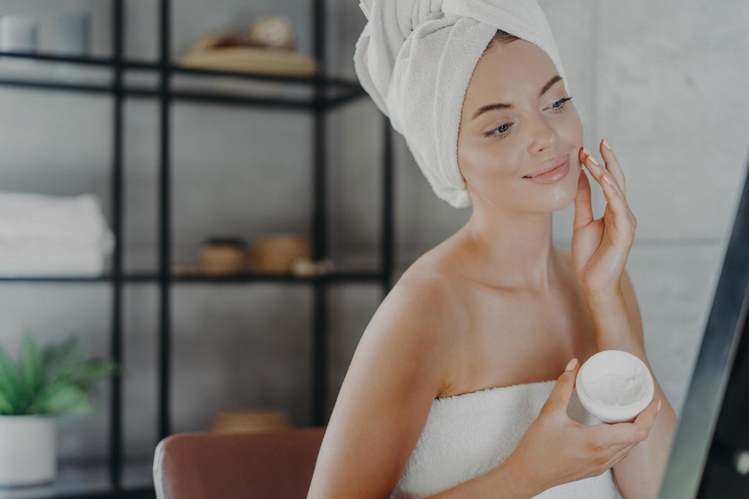 une jeune femme européenne ravie met de la crème sur le visage, regarde son reflet dans le miroir, enveloppée dans une serviette de bain, a une procédure anti-âge après la douche pour une peau saine. concept de soins de la peau photo