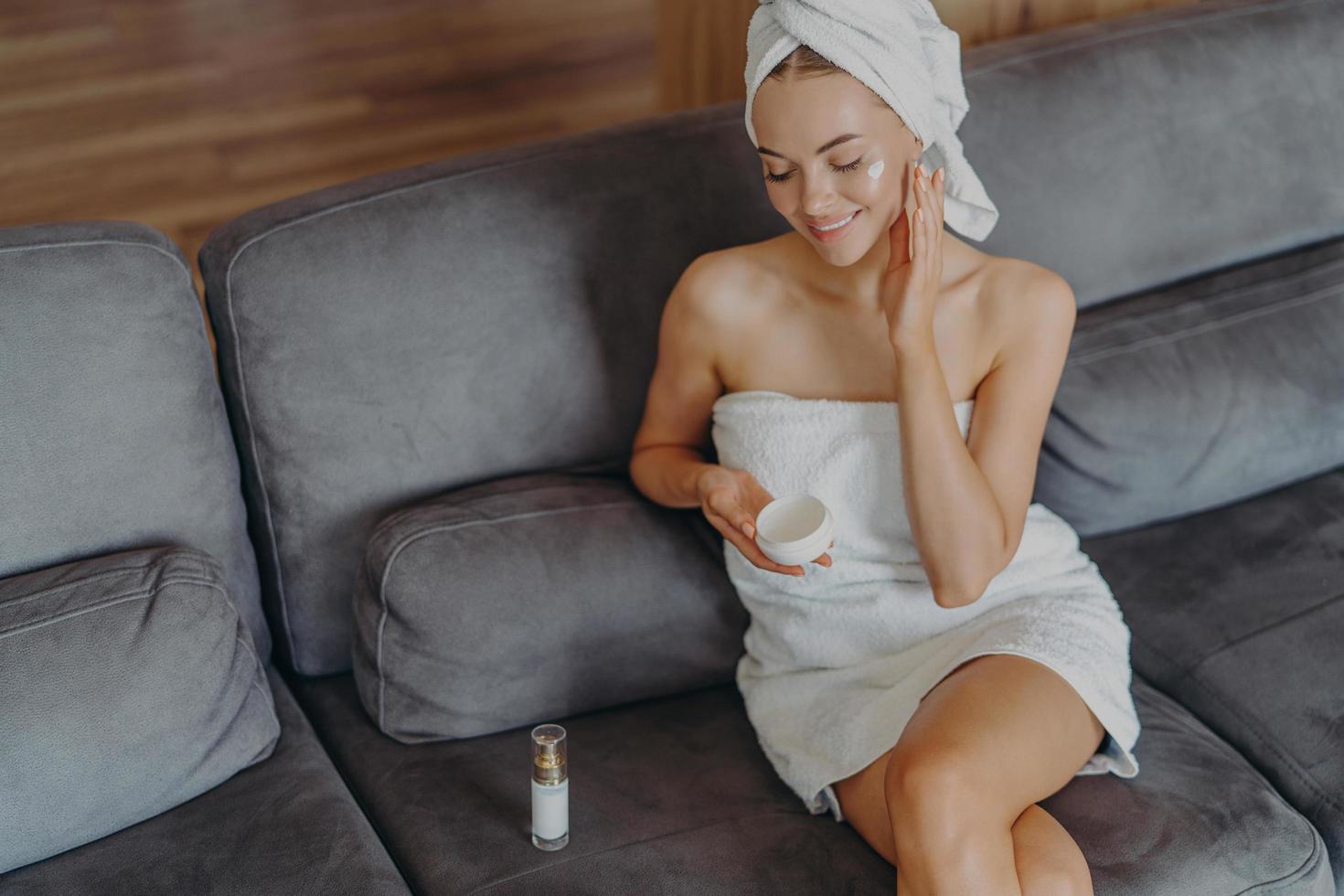 vue de dessus d'une femme heureuse en bonne santé assise sur un canapé confortable à l'intérieur, applique de la crème pour le visage et utilise une lotion pour le corps après avoir pris un bain, a un maquillage naturel et une peau saine. concept d'hygiène et de beauté photo