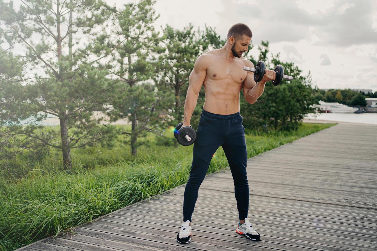 concept de sport et de musculation. un homme européen barbu musclé soulève des haltères lourdes porte un pantalon de sport et des baskets fléchit les muscles à l'extérieur, fait des exercices d'haltérophilie, mène une vie active photo