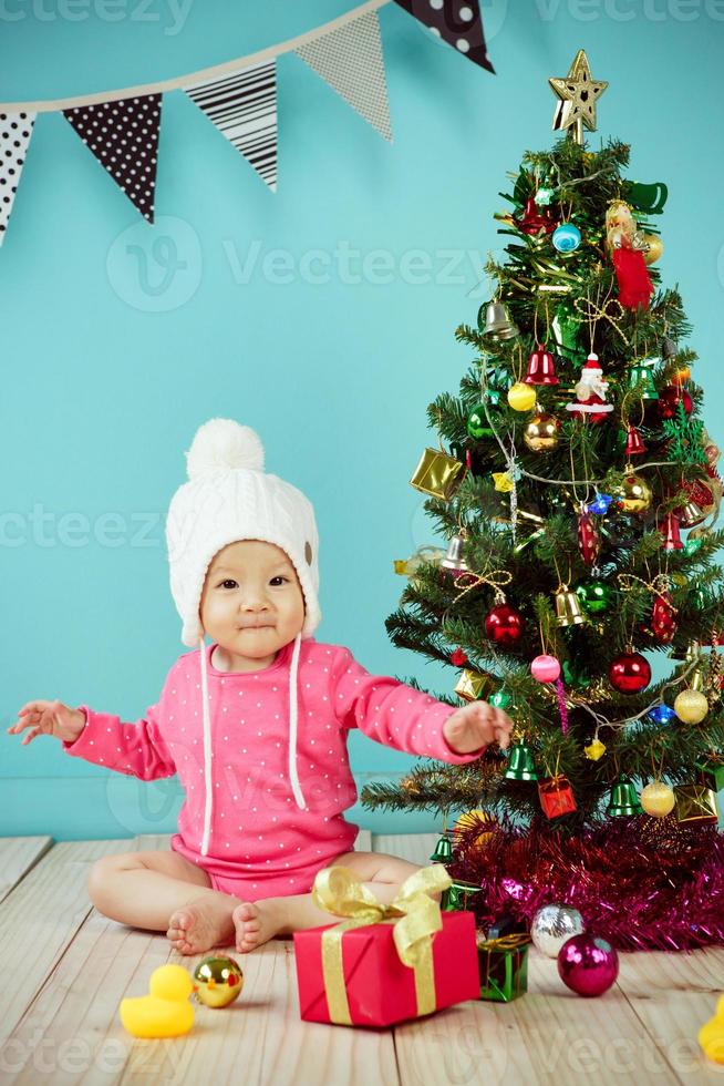 bébé portant un bonnet tricoté blanc devant un fond bleu et décorant un sapin de noël sur fond vert photo