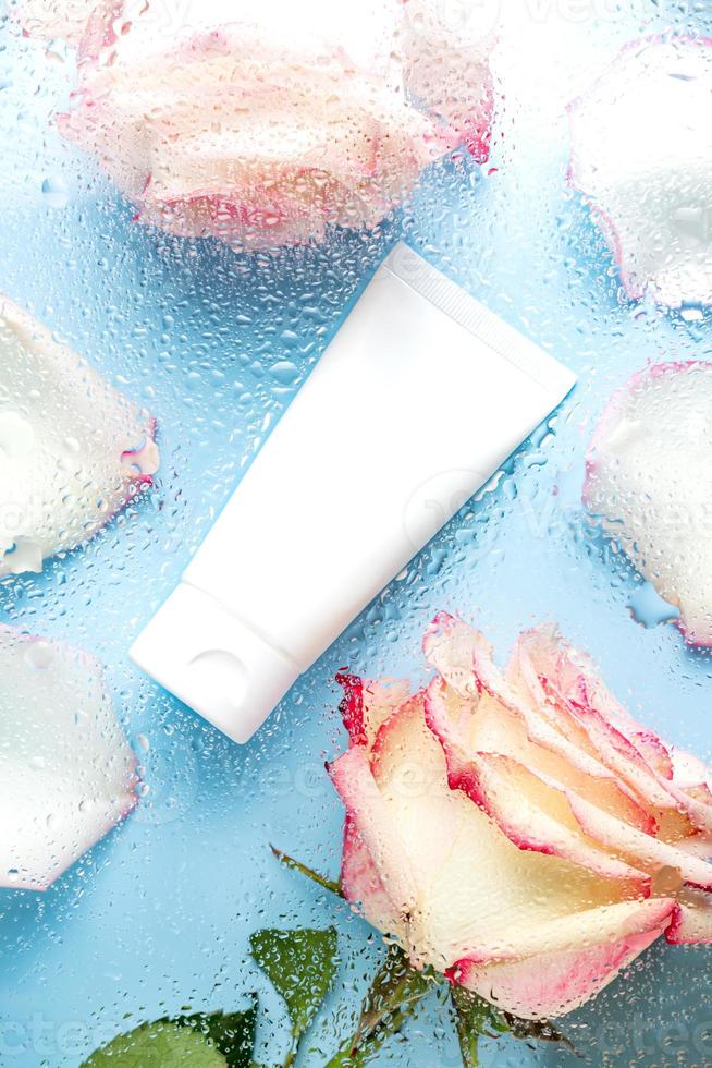 tube cosmétique pour crème pour le visage, nettoyant ou lotion pour le corps sur fond bleu avec des gouttelettes d'eau et des fleurs roses à travers le verre. le concept d'un produit cosmétique hydratant photo