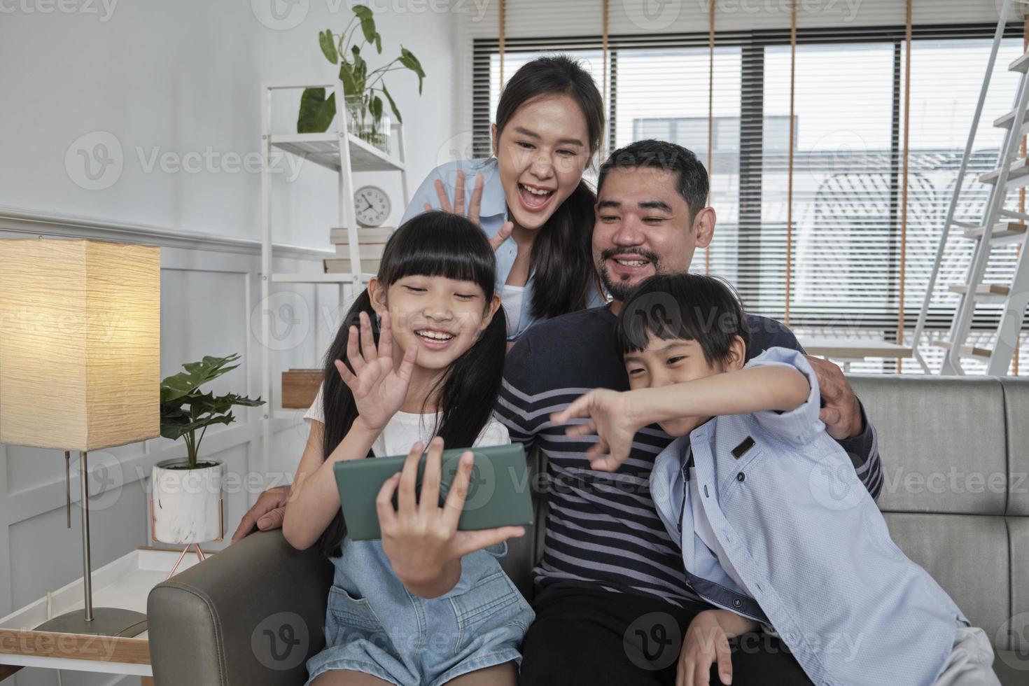 heureux parents thaïlandais asiatiques et enfants appel vidéo en ligne avec la famille via Internet sur le canapé dans le salon de la maison, beau week-end et style de vie de bien-être domestique avec la technologie Internet téléphone mobile. photo