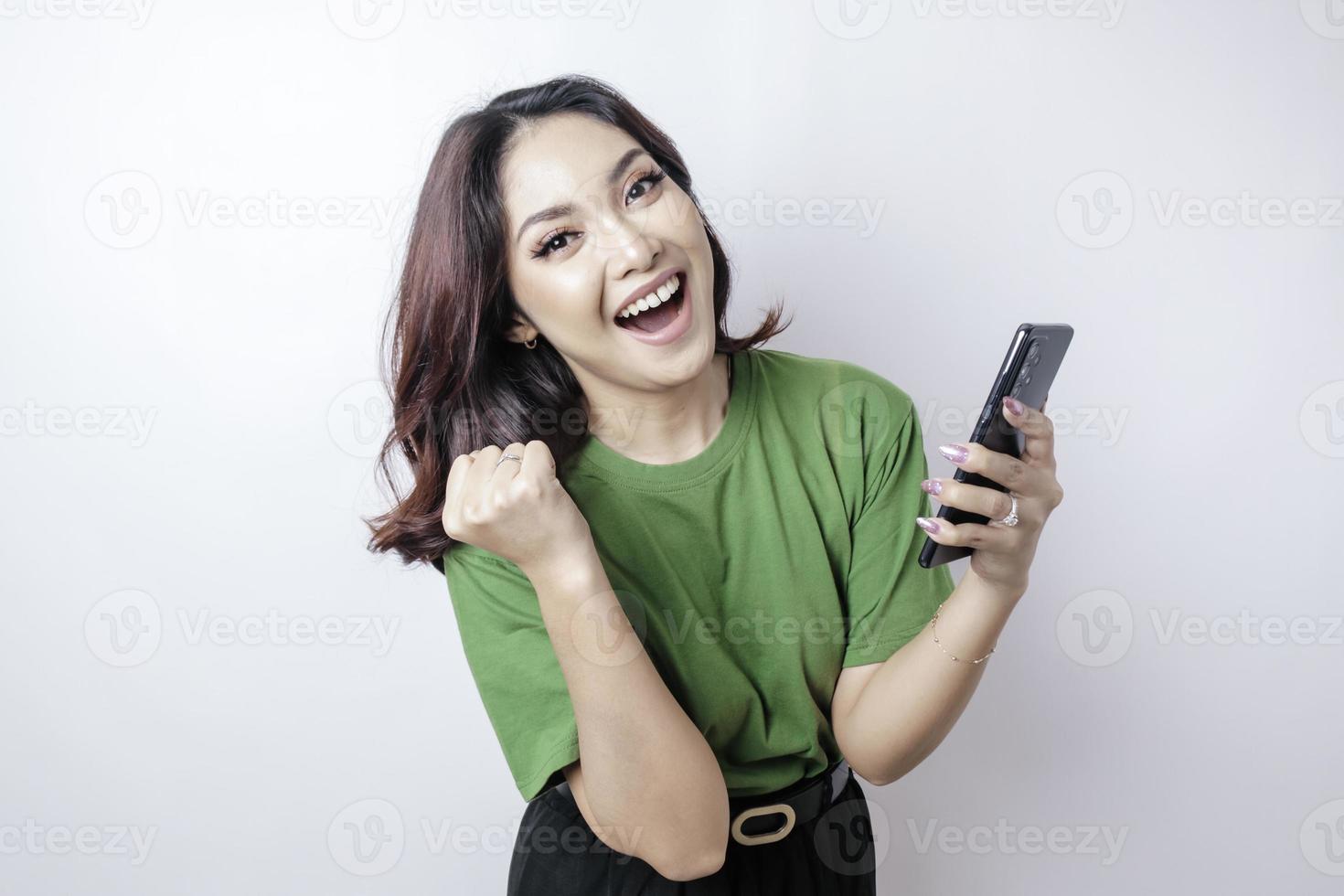 une jeune femme asiatique avec une expression heureuse et réussie portant un t-shirt vert et tenant un smartphone isolé sur fond blanc photo