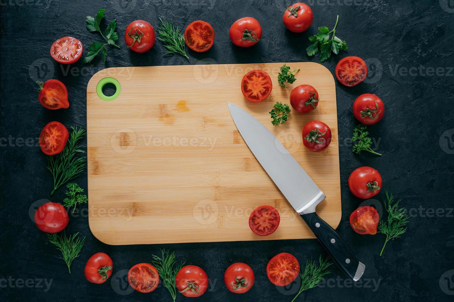 cadre de tomates autour d'une planche à découper en bois. légumes mûrs et tranches, persil vert et aneth près de la planche de cuisine et du couteau. art culinaire photo