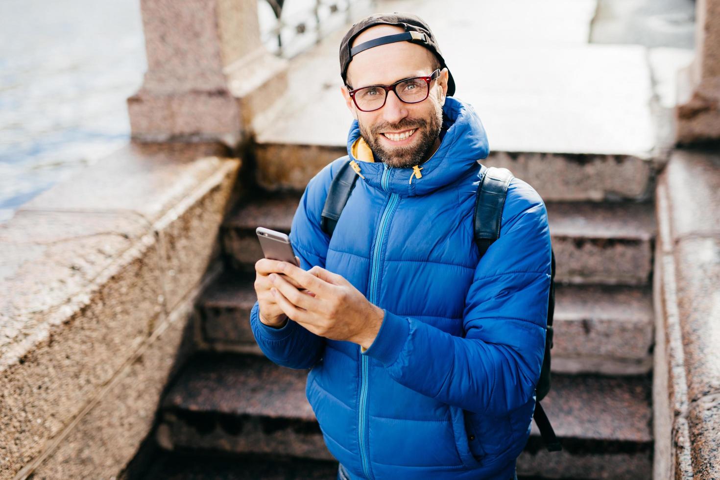 jeune homme barbu en casquette noire et anorak bleu tenant un sac à dos debout à l'extérieur avec un téléphone portable isolé sur fond d'étapes. blogueur élégant avec smartphone. concept de personnes et de style de vie photo