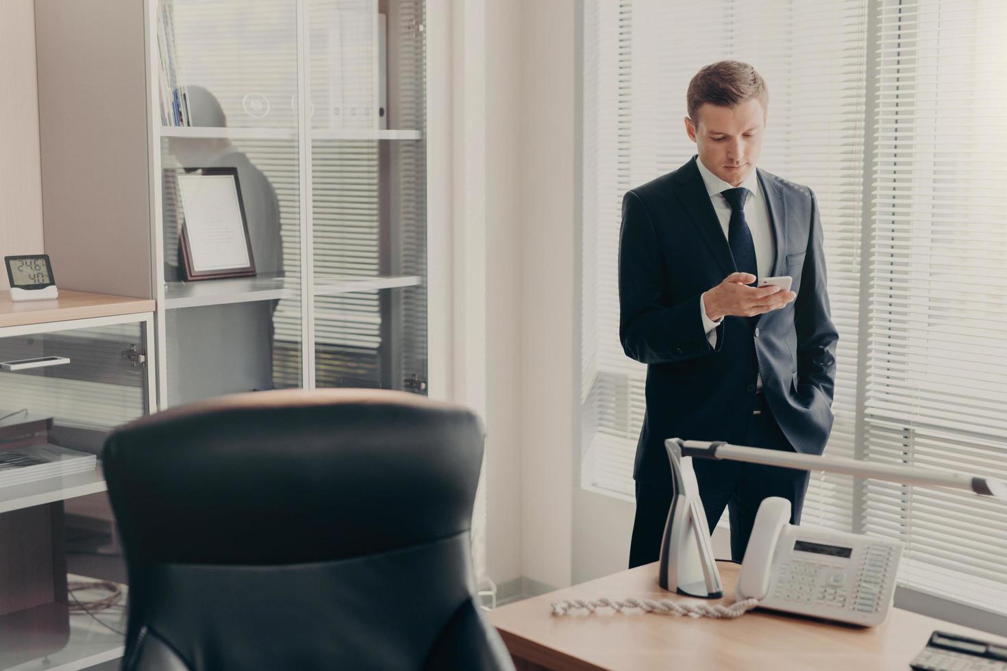 beau jeune directeur masculin en vêtements d'entreprise, navigue sur des pages Web et discute en ligne avec un smartphone, a un look concentré, pose dans un intérieur de bureau moderne, lit les actualités et parcourt les informations photo