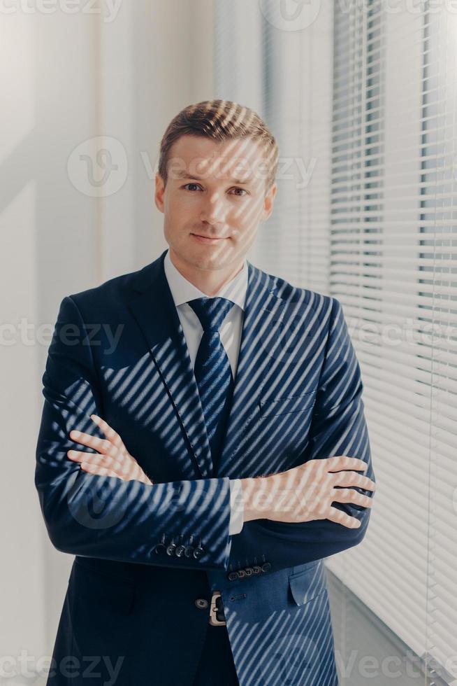 photo verticale d'un directeur masculin sérieux qui garde les mains croisées, vêtu d'un élégant costume noir, se tient près de la fenêtre du bureau dans son cabinet, profite d'une atmosphère calme après une journée de travail, pense à quelque chose