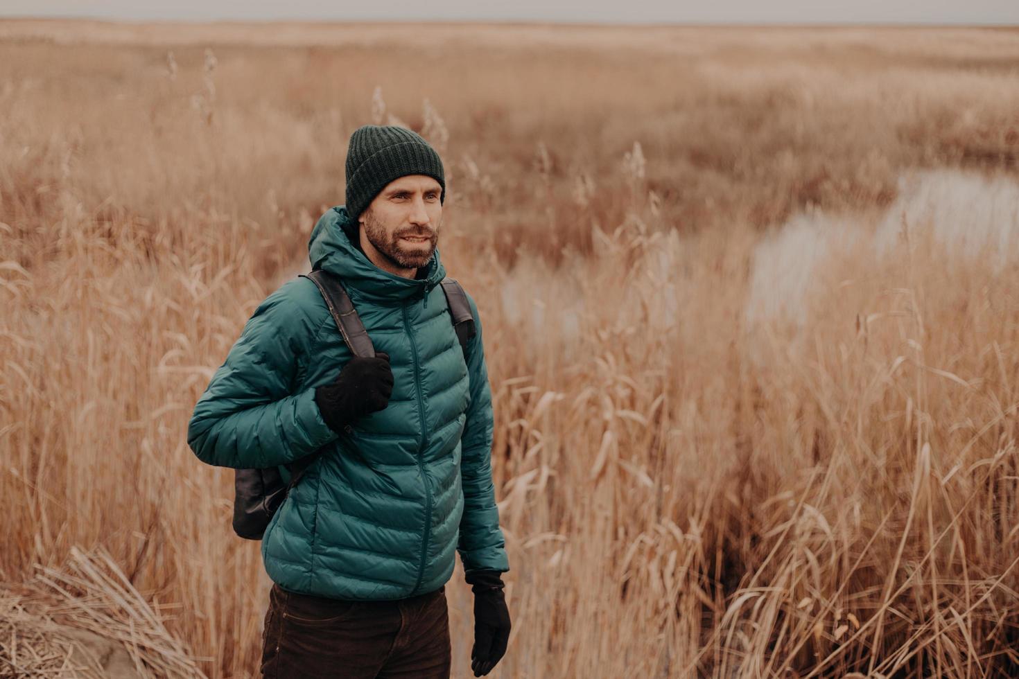 photo en extérieur d'un voyageur masculin pensif vêtu de vêtements chauds, de bonne humeur, portant un sac à dos, se promenant dans un cadre rural près du champ en automne, profitant d'une atmosphère calme. notion de loisirs