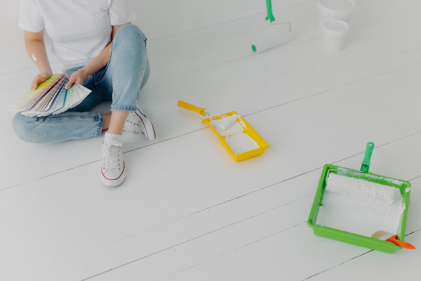 image recadrée d'une femme sans visage choisit la couleur à partir d'échantillons, va remettre à neuf les murs de sa propre chambre, porte des baskets, s'assoit sur un mur blanc avec des plateaux et des rouleaux de peinture. concept de réparation et de conception photo