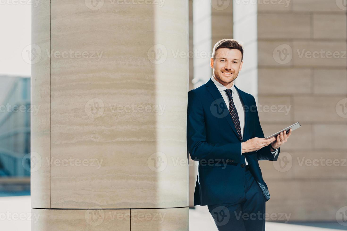photo horizontale d'un directeur masculin prospère ravi et heureux utilise un ordinateur tablette moderne, vêtu d'un costume formel, se tient près d'un immeuble de bureaux, a une expression confiante et heureuse. employé de bureau avec appareil