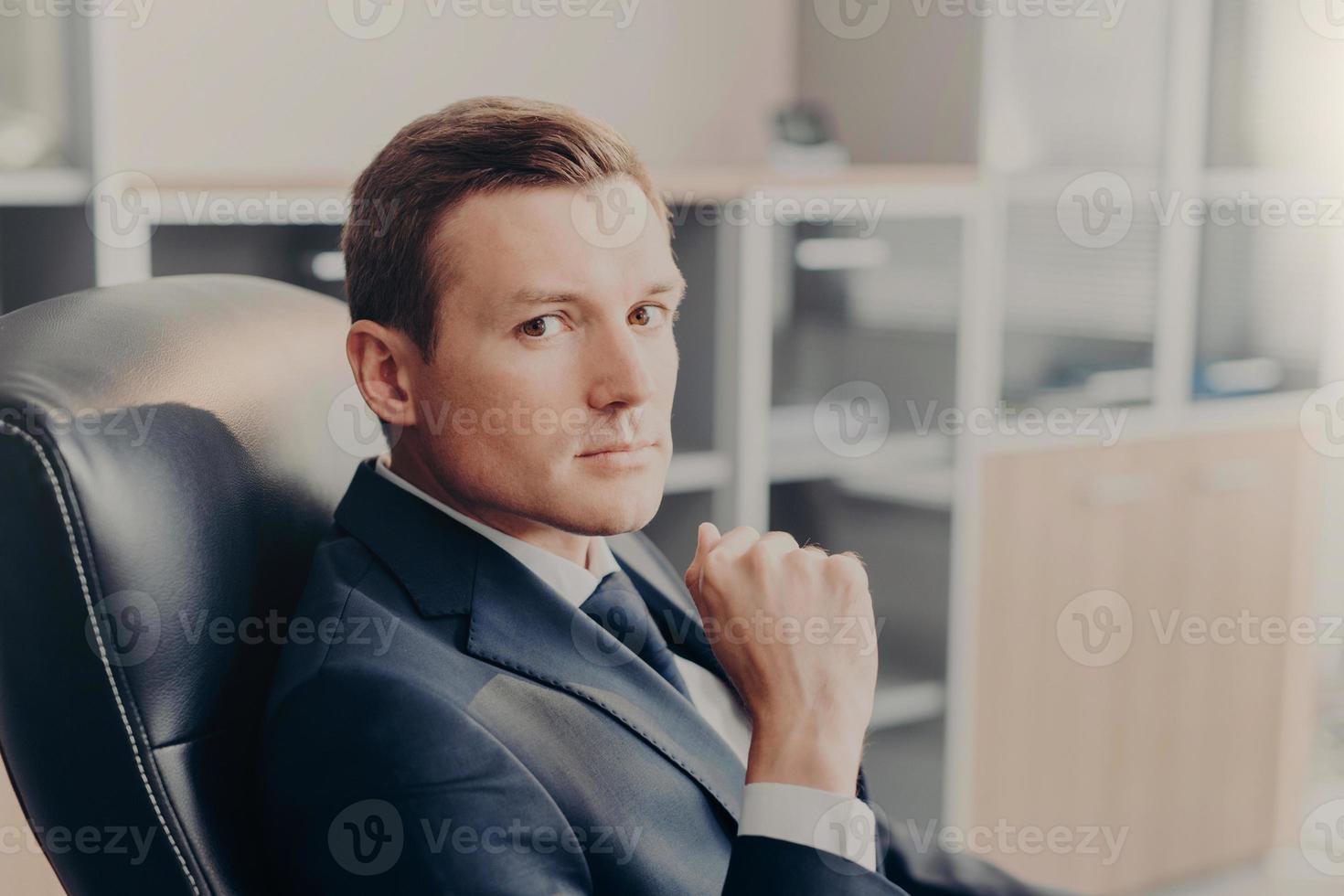 directeur masculin professionnel en costume formel, regarde directement la caméra, se repose sur une chaise sur le lieu de travail, envisage un nouveau projet d'entreprise. directeur homme élégant travaille dans sa propre entreprise ou entreprise photo