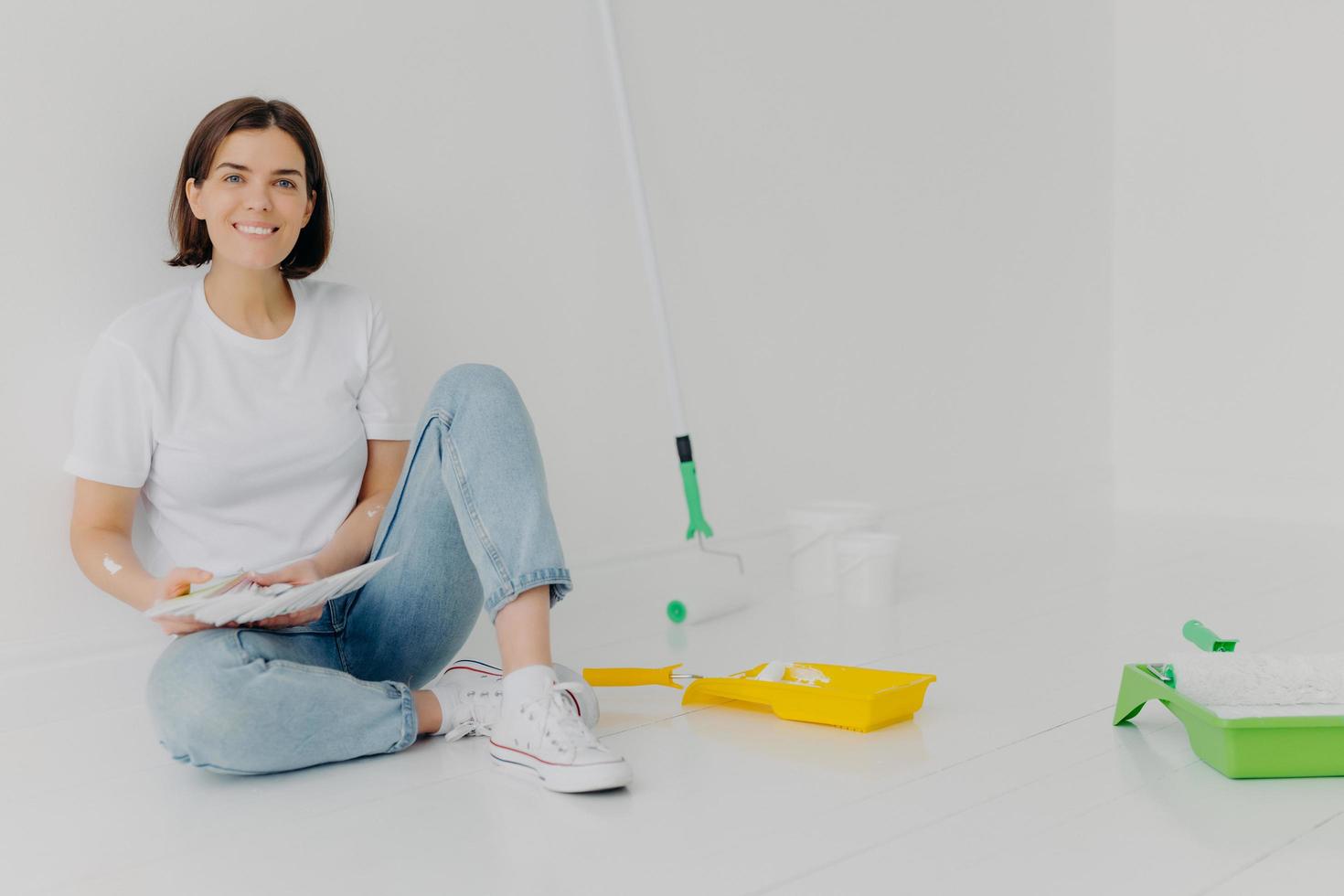 photo horizontale d'une designer souriante qui choisit la meilleure couleur pour l'appartement, tient la palette, vêtue de vêtements décontractés, peint les murs, rénove sa maison, réfléchit à des idées créatives pour décorer la chambre