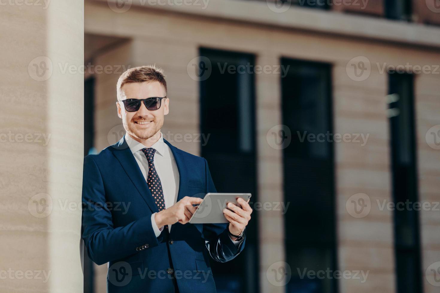 un propriétaire d'entreprise élégant portant des lunettes de soleil à la mode utilise une tablette numérique moderne pour participer à une conférence en ligne avec des partenaires commerciaux, a un sourire positif sur le visage, connecté à Internet sans fil photo