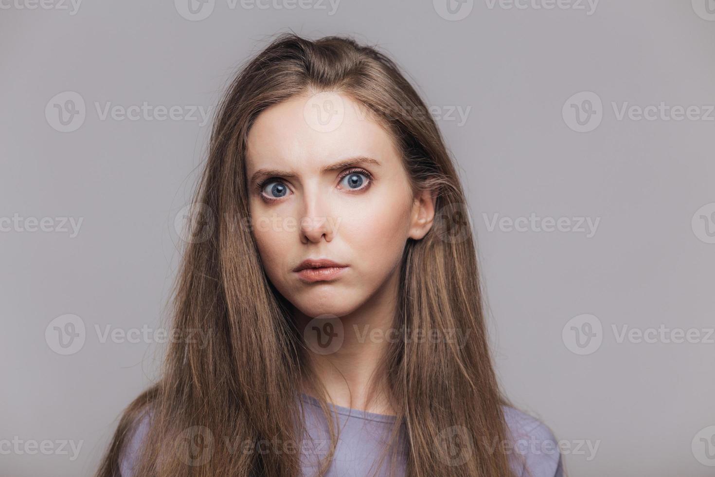 photo de tête d'une belle femme aux yeux bleus et à la peau pure, regarde sérieusement et avec colère la caméra, a les cheveux raides foncés, isolée sur fond gris. photo horizontale d'une belle jeune femme à l'intérieur