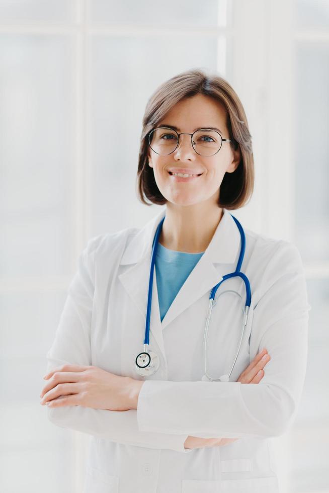 belle femme médecin joyeuse porte une robe blanche, des lunettes et un phonendoscope, garde les mains croisées, regarde avec confiance la caméra, a les cheveux noirs courts, se dresse sur fond blanc, travaille en clinique photo