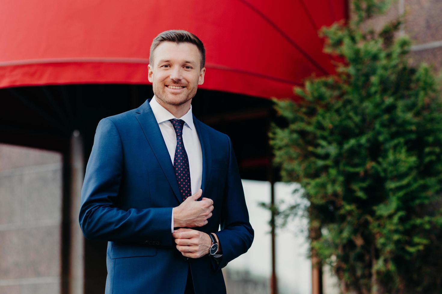 un cadre masculin positif porte un costume noir formel, a une expression positive, se tient à l'extérieur, attend quelqu'un, est confiant dans la réussite de son travail acharné et de sa persévérance photo