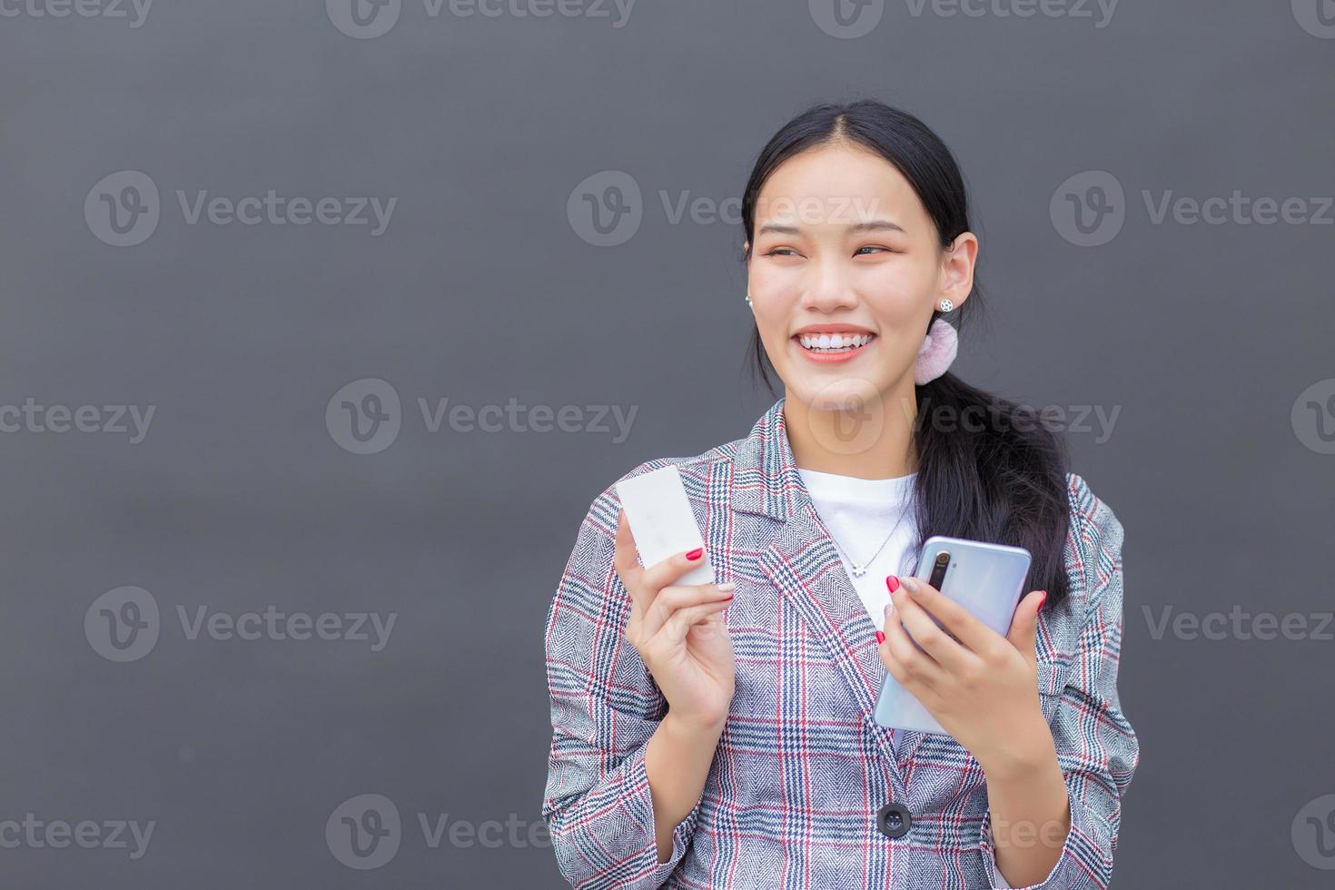 une femme asiatique professionnelle dans un blazer à rayures marron sourit joyeusement, réussie et confiante tout en tenant un smartphone et une carte de crédit sur le mur gris foncé. photo
