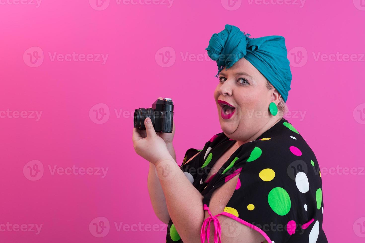 femme souriante de grande taille avec une drôle d'expression de visage émotionnel avec appareil photo vintage en robe denim isolée sur fond rose, voyageur en vacances, style de mode d'été, touriste excité.