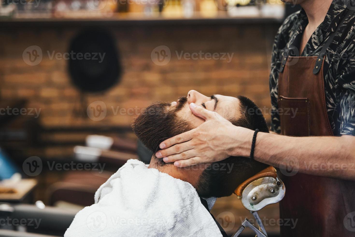 bel homme avec une barbe et les yeux fermés dans une cape de coupe de cheveux noire dans le salon de coiffure. barbier lui fait un massage du visage. fermer. horizontal. photo