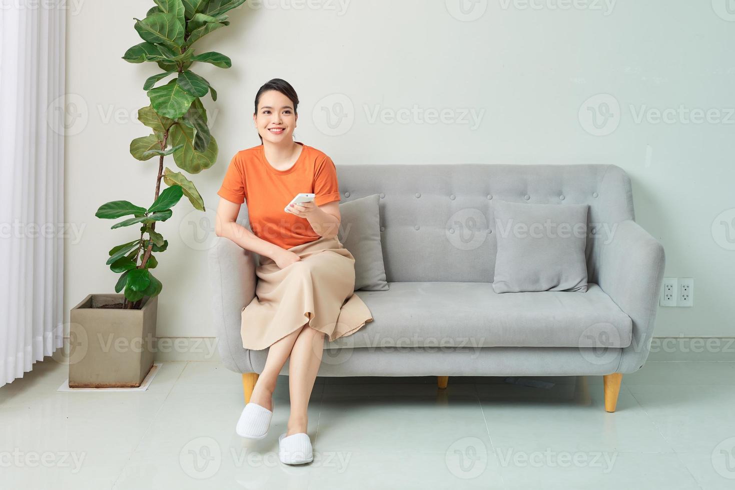 belle femme souriante à l'aide de la télécommande du climatiseur assis sur un canapé à la maison photo