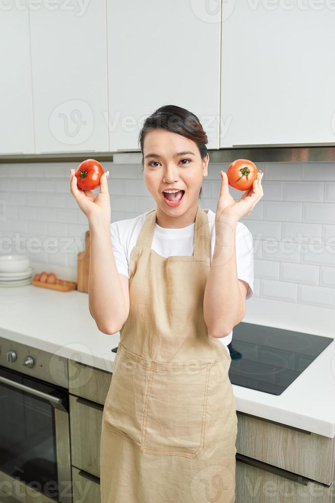 attrayant drôle femme au foyer dame bras tenant deux grosse tomate humeur enjouée profiter matin cuisine savoureux dîner attendre famille invités porter tablier stand cuisine moderne à l'intérieur photo