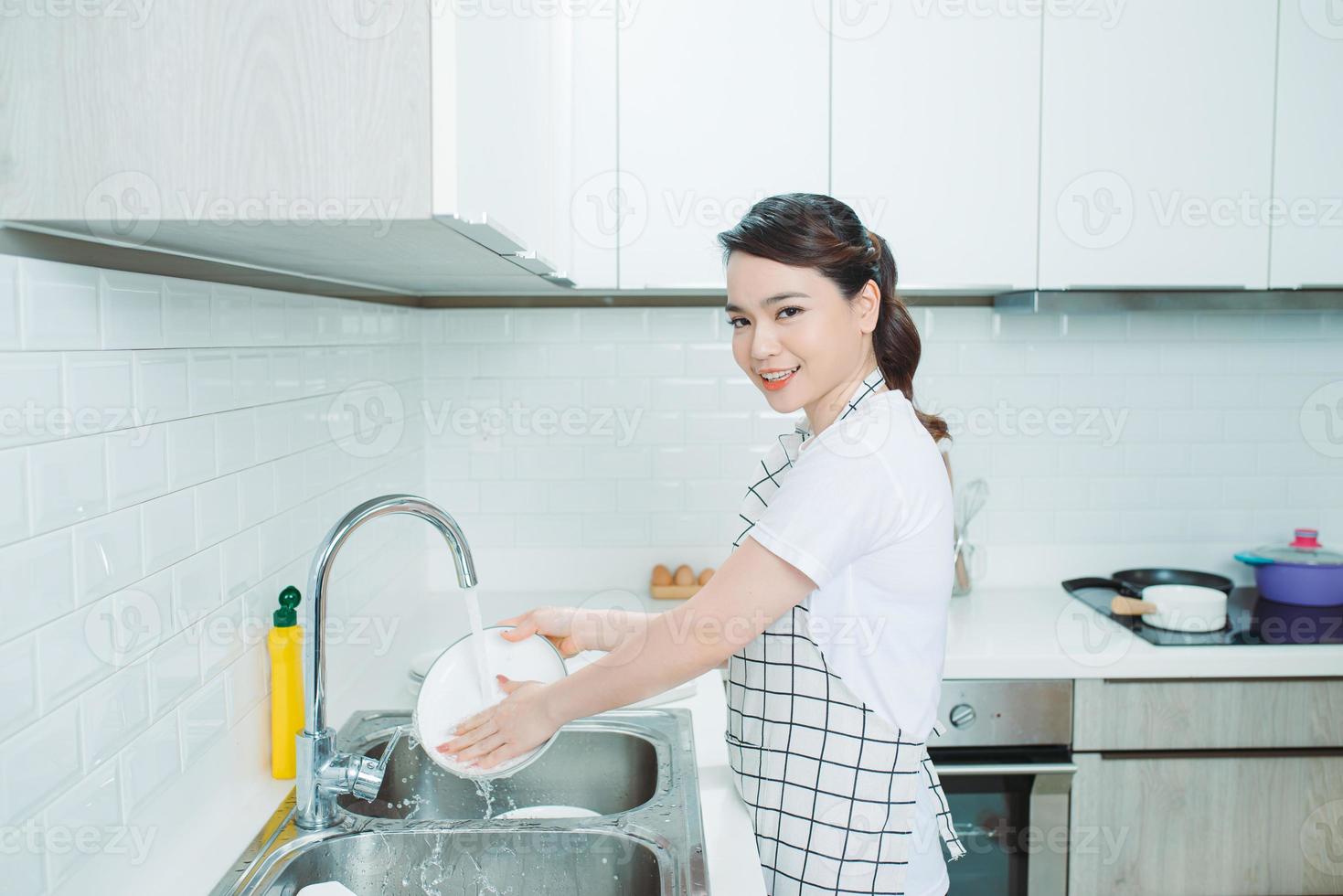 belle jeune femme souriante faisant la vaisselle dans une cuisine blanche moderne. photo