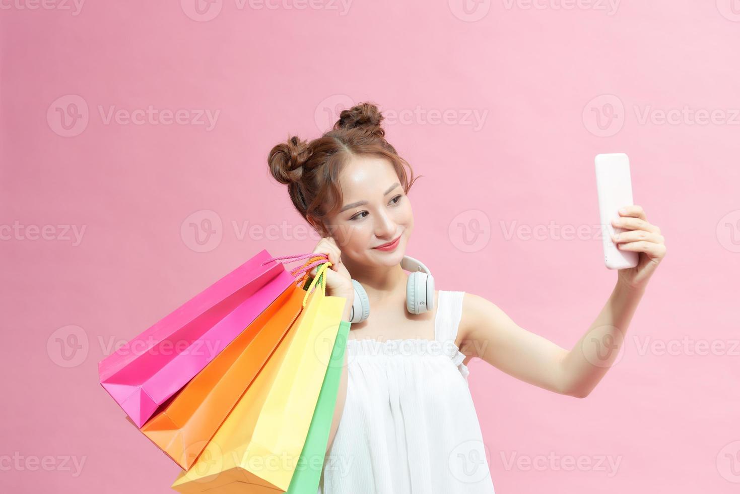 belle jeune femme asiatique heureuse tenant un smartphone pour selfie et faire du shopping en ligne avec des sacs à provisions sur fond rose. photo