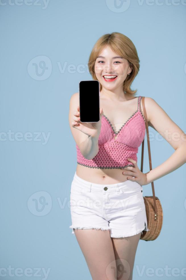 femme asiatique heureuse tenant et montrant un smartphone innovant riant de la caméra sur fond bleu photo