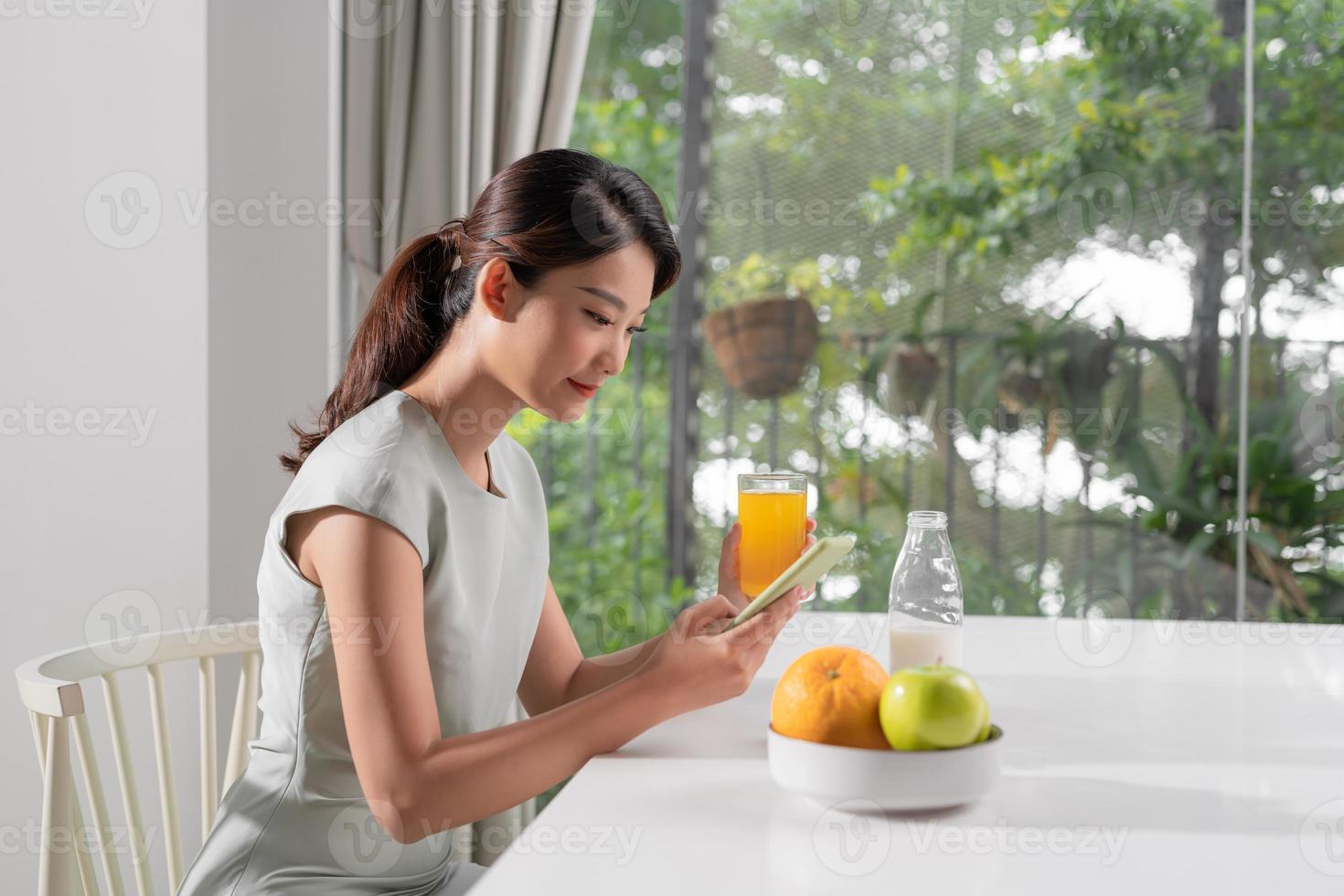 jolie femme souriante regardant un téléphone portable et tenant un verre de jus d'orange tout en prenant son petit déjeuner photo