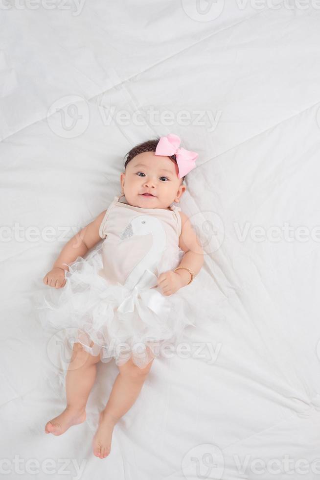 Smiling baby girl toddler in white dress and hair bow decoration lying on bed photo