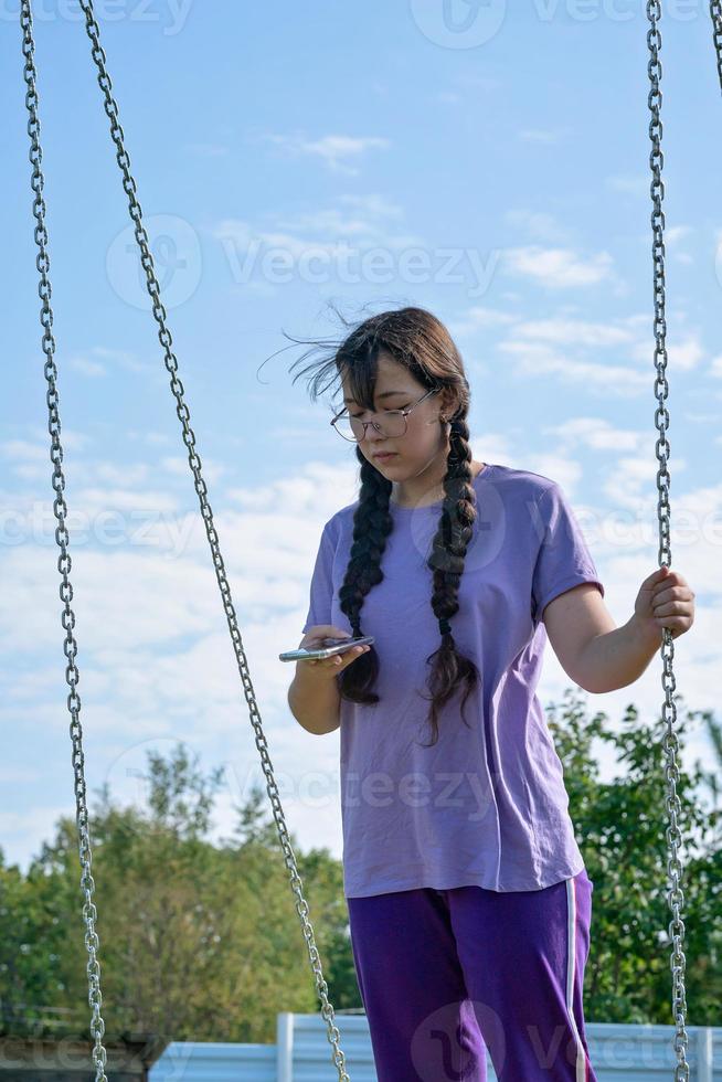 une adolescente se balance sur une balançoire avec un téléphone dans les mains, communique sur les réseaux sociaux ou apprend en ligne photo