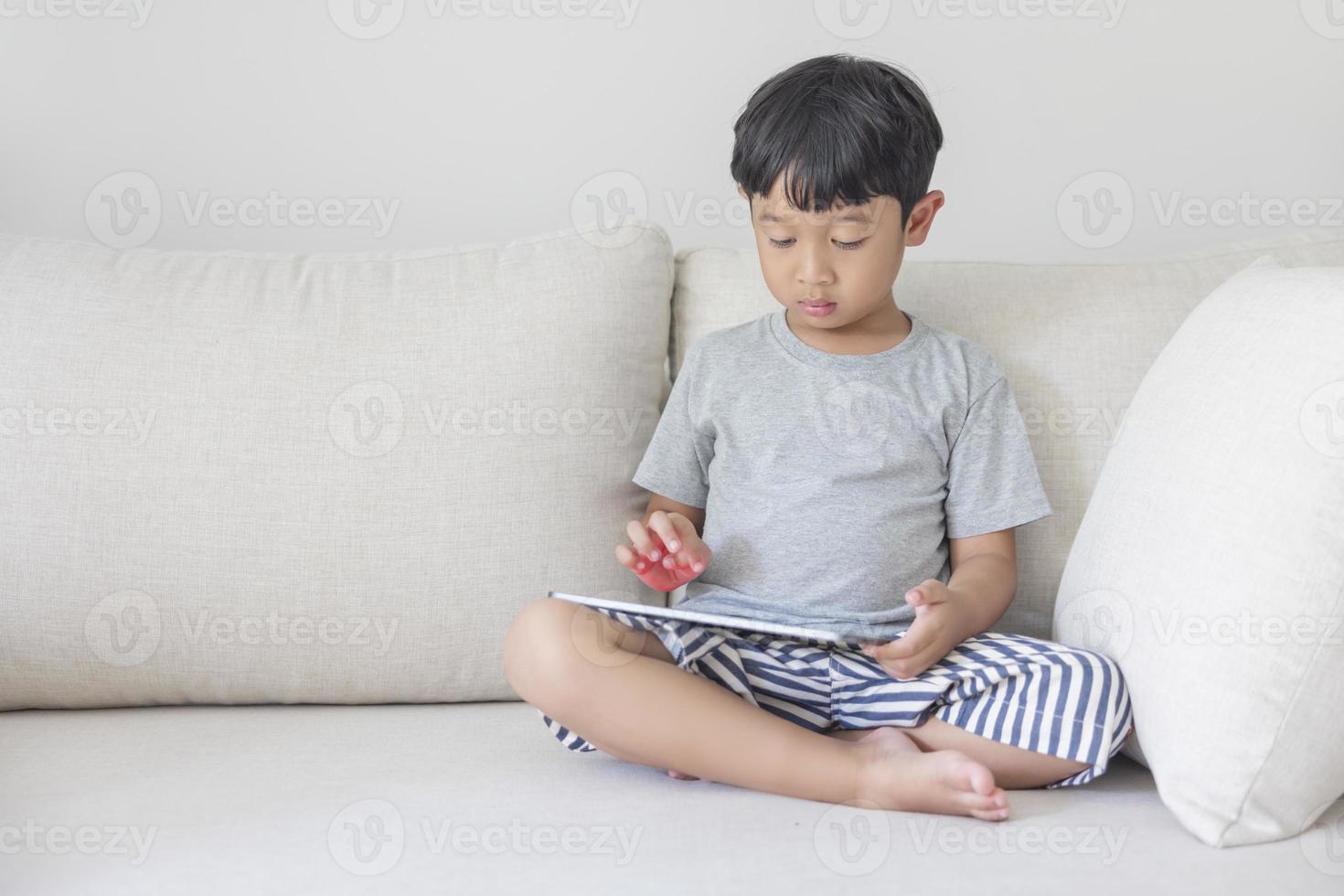 adorable garçon asiatique heureux vêtu d'une chemise grise et d'un short rayé bleu-blanc s'amuse à jouer avec sa tablette sur un canapé crème. regardant l'écran du mobile photo