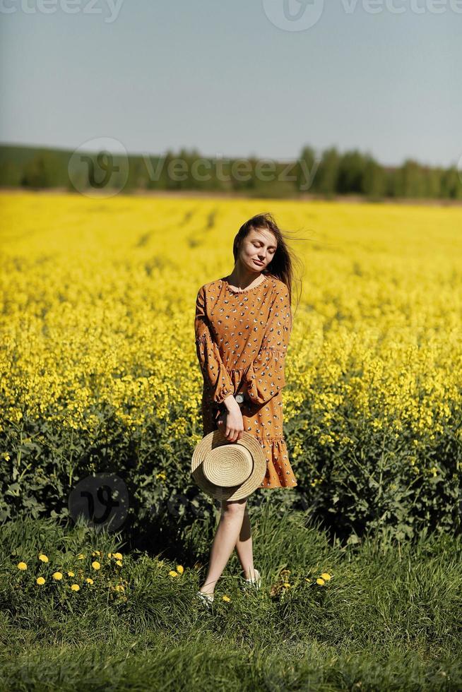 belle jeune femme se promène dans un champ de colza jaune. fille brune cheveux longs voler dans le vent vêtue d'une robe et d'un chapeau de paille. notion de vacances d'été photo