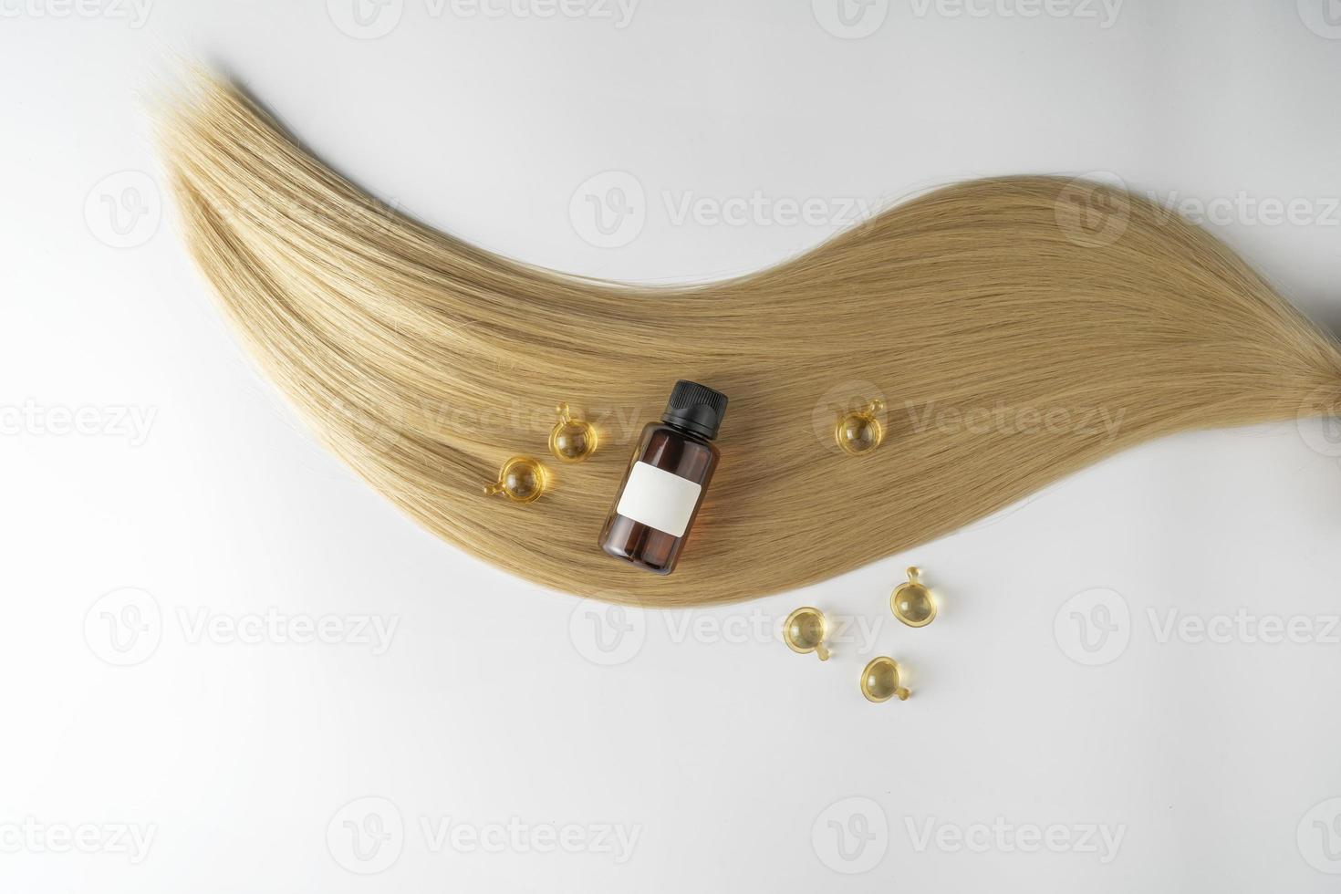 une huile ou un sérum de soin des cheveux dans un flacon compte-gouttes marron et des capsules dorées posées sur une mèche de cheveux blonds, maquette de marketing de produit photo