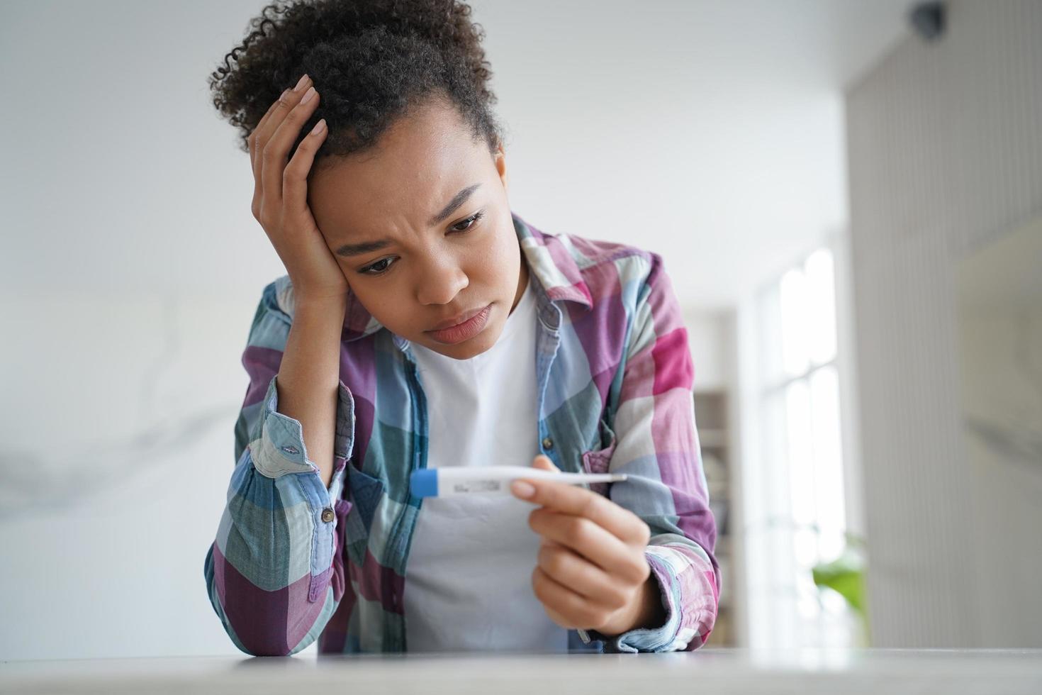 une fille métisse malsaine tient un thermomètre souffre d'une température corporelle élevée, de fièvre, de symptômes covid photo