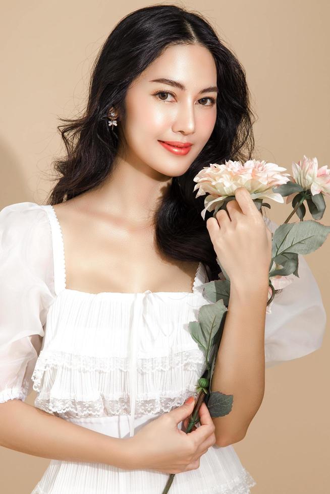 femme asiatique avec un beau visage et une peau fraîche parfaite et propre avec une fleur. modèle féminin mignon avec maquillage naturel et yeux pétillants sur fond beige isolé. photo