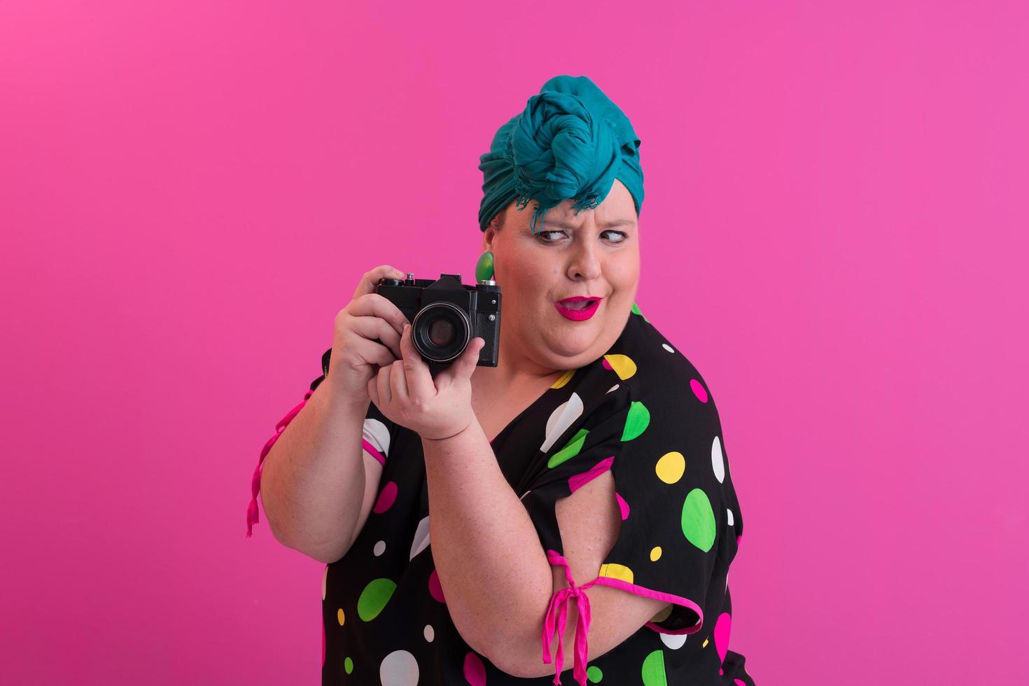 femme souriante de grande taille avec une drôle d'expression de visage émotionnel avec appareil photo vintage en robe denim isolée sur fond rose, voyageur en vacances, style de mode d'été, touriste excité.