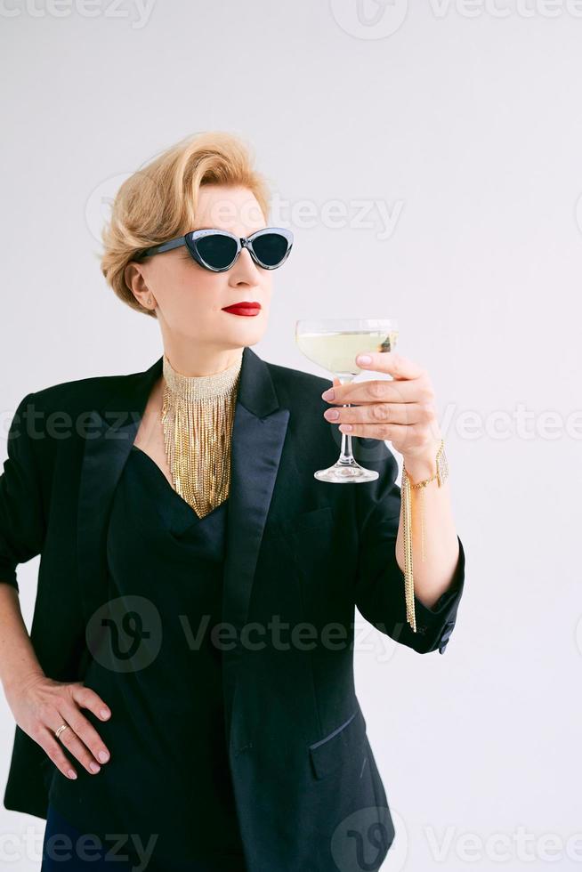 femme élégante et élégante mature en smoking et lunettes de soleil avec un verre de vin mousseux. fête, célébration, concept anti-âge photo