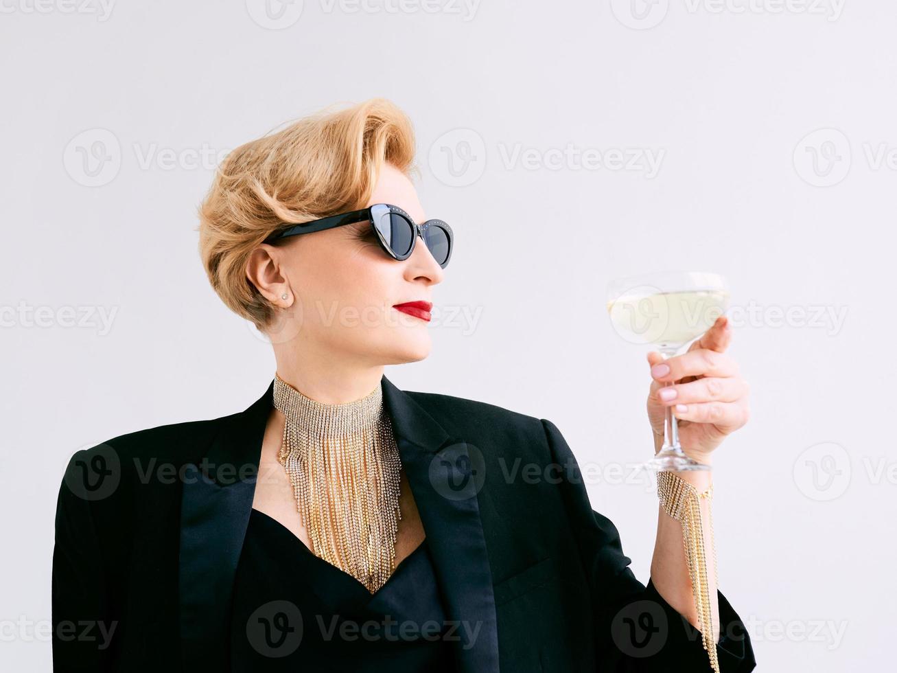 femme élégante et élégante mature en smoking et lunettes de soleil avec un verre de vin mousseux. fête, célébration, concept anti-âge photo