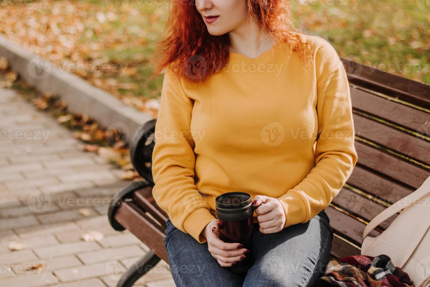portrait de jeune femme rousse assise dans le parc sur un banc avec une tasse de café. la dame porte un sweat-shirt jaune et tient une tasse réutilisable. mode de vie le jour d'automne ensoleillé. photo