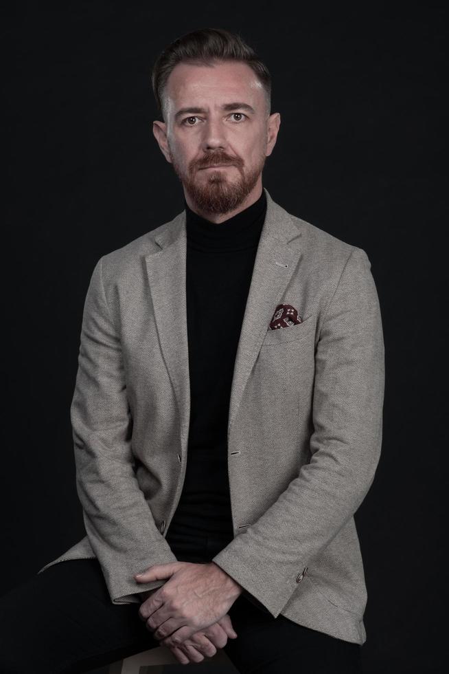 portrait d'un homme d'affaires adulte portant un costume à la mode et assis dans un studio moderne sur une chaise élégante sur fond noir photo